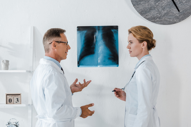 doctor in glasses gesturing near x-ray while looking at attractive coworker  - Photo, Image