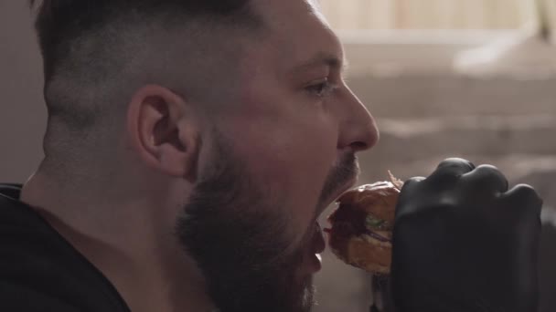 Side portrait of a bearded man in black gloves eating a tasty burger. The man enjoying mouth-watering fast food in the modern restaurant. - Záběry, video