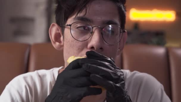 Close-up portrait of cute skinny young man in glasses and black gloves eating tasty shawarma. The man enjoying mouth-watering fast food in the modern restaurant. - 映像、動画