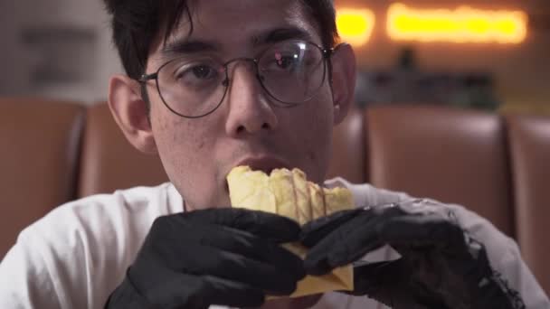 Close-up portrait of a skinny young man in glasses and black gloves eating tasty shawarma. The man enjoying mouth-watering fast food in the modern restaurant - Séquence, vidéo
