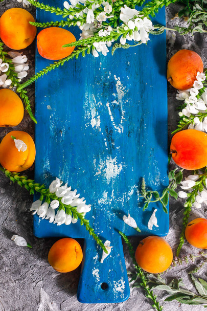 Friss sárgabarack és kék fából készült tábla konkrét hátteret friss virágokkal. A kajszibarack nyári betakarításának összetétele. - Fotó, kép