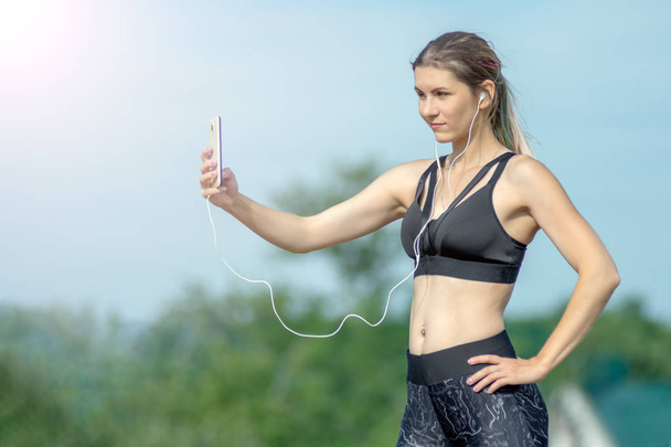 Chica sexy deportiva de fitness haciendo foto selfie en el teléfono inteligente durante el descanso de entrenamiento. Parque de la ciudad y cielo azul en el fondo
 - Foto, Imagen