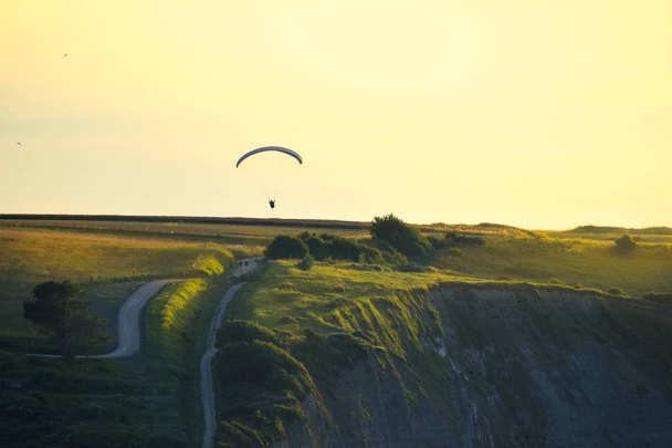 Rotsachtige kliffen bedekt met groen gras bij zonsondergang. Een man oefent paragliding over hen. Normandië, Frankrijk. - Foto, afbeelding