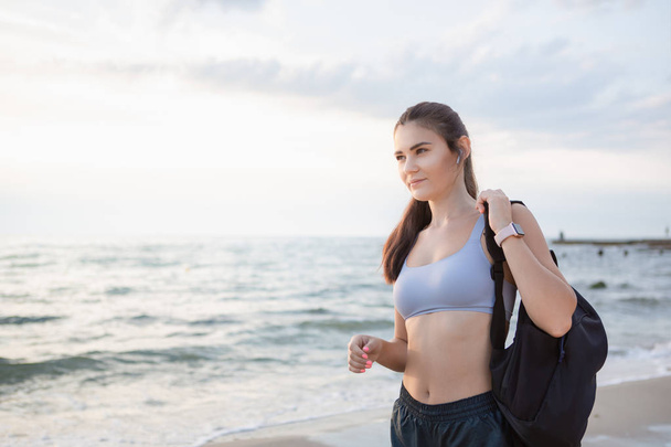 ワイヤレスイヤホン、スマートウォッチ、黒いバックパックを持つ若いブルネットの女性は、日の出の海岸で朝のトレーニングの後に休む. - 写真・画像