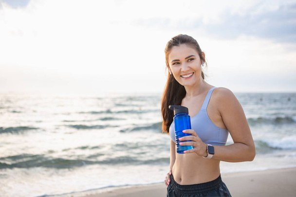 Young brunette woman with wireless earbuds and smart watches resting after morning workout at the sea shore at sunrise drinking water - Foto, Imagem