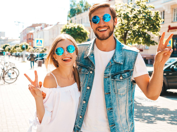 Smiling beautiful girl and her handsome boyfriend. Woman in casual summer dress.Man in jeans clothes. Happy cheerful family. Female having fun on the street background.Shows peace sign in sunglasses - Photo, image