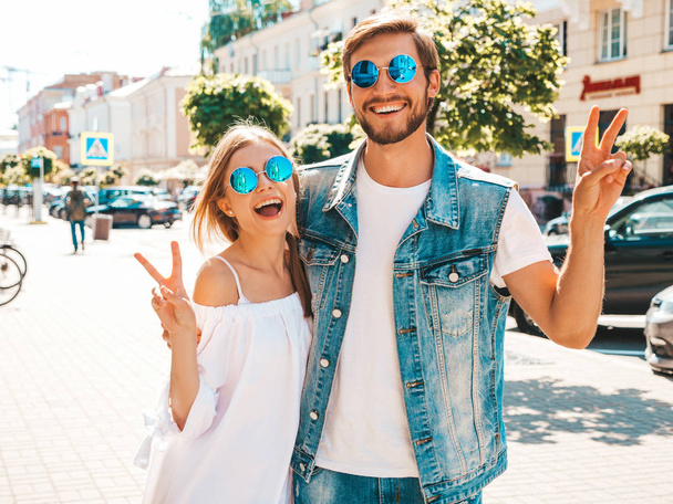 Smiling beautiful girl and her handsome boyfriend. Woman in casual summer dress.Man in jeans clothes. Happy cheerful family. Female having fun on the street background.Shows peace sign in sunglasses - Zdjęcie, obraz