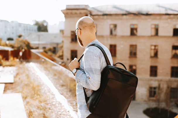 Красивий хлопець зі стильним рюкзаком, дивиться вбік, відповідає стоячи на терасі, розмитий будівельний фон
 - Фото, зображення