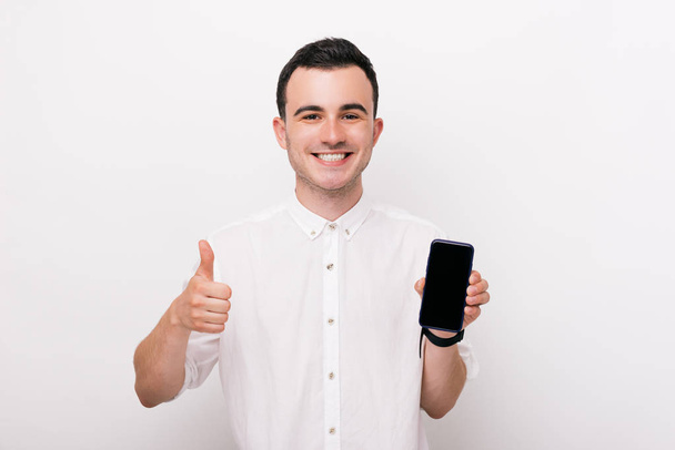 Photo of handsome young man, wearing a white shirt, holding a smartphone and showing thumb up - Фото, изображение