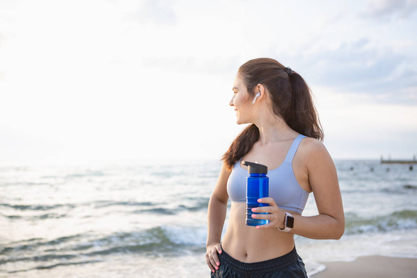Young brunette woman with wireless earbuds and smart watches resting after morning workout at the sea shore at sunrise drinking water - Fotoğraf, Görsel