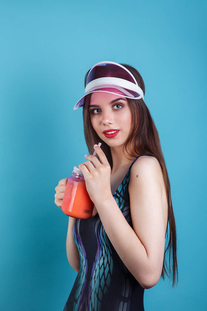 Sexy woman in swimsuit and plastic visor holding glass of fresh summer drink on blue background - Φωτογραφία, εικόνα
