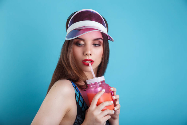 Sexy woman in swimsuit and plastic visor holding glass of fresh summer drink on blue background - Fotó, kép