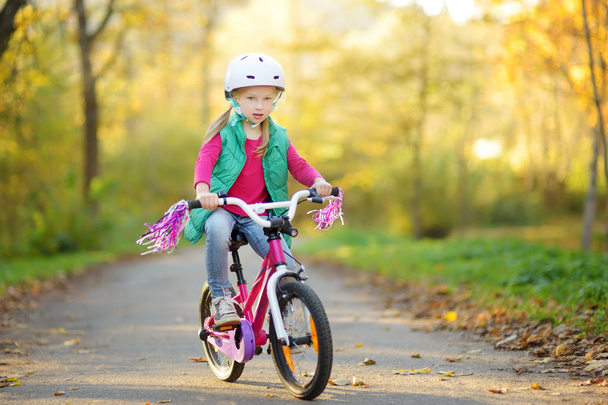Cute Little Girl jazda na rowerze w parku miejskim w słoneczny jesienny dzień. Aktywny wypoczynek rodzinny z dziećmi. - Zdjęcie, obraz