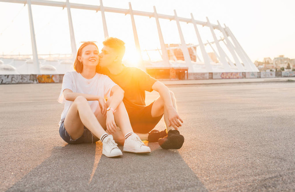 Любляча пара сидить на вулиці на фоні заходу сонця, хлопець цілує дівчину на щоці, дівчина дивиться на камеру і посміхається. Пара відпочиває на вулиці в теплий літній вечір
 - Фото, зображення