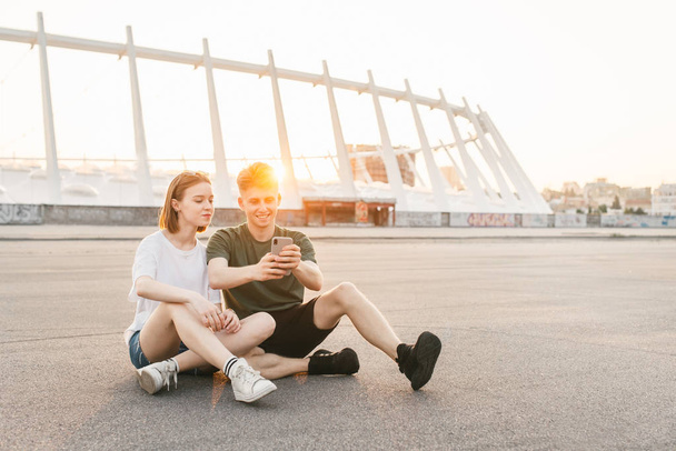 Couple moderne de milenials assis dans la rue en arrière-plan d'un beau paysage urbain au coucher du soleil, à l'aide d'un smartphone et souriant. Étudiants assis sur un beau fond urbain et se détendre
. - Photo, image