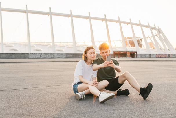 Cute para w stylowych ubrań siedzi na chodniku, piękny krajobraz miejski o zachodzie słońca i za pomocą smartfona. Młody człowiek i dziewczyna odpocząć na ulicy, siedzieć na ziemi i zrobić zdjęcie. - Zdjęcie, obraz