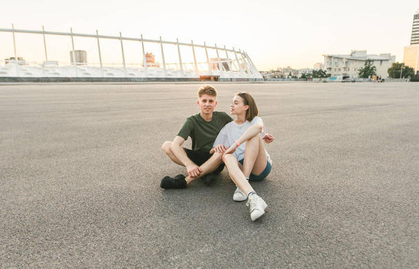 Bella coppia seduta sul marciapiede e abbracciata sullo sfondo del tramonto e del paesaggio urbano.Coppia carina in abiti eleganti che si riposa su una passeggiata, si siede in piazza e guarda nella fotocamera
 - Foto, immagini