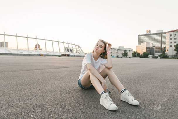 Attractive girl sits in the background of the city landscape and relaxes, wears a stylish casual clothing and looks at the camera. . The act in a white T-shirt and sneakers sits on the asphalt and pos - 写真・画像