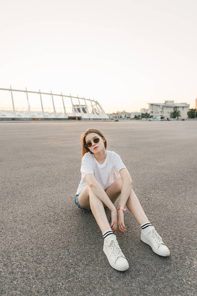 Trendy street portrait of a sexy girl in a stylish casual clothing and sunglasses sits on the street and looks at camera,background of asphalt. Evening lifestyle portrait of a girl.Vertical photo - Fotó, kép