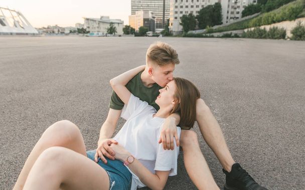 愛するカップルは、街の風景を背景に舗装に横たわって抱擁し、キス。若い男は額にガールフレンドにキスし、女の子は微笑みます。ラブストーリー。ストリート写真. - 写真・画像