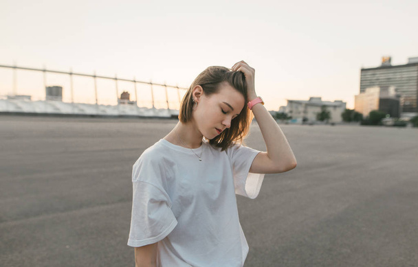 Portrait of an unhappy girl standing with her eyes closed against the backdrop of a city landscape in the sunset and fixing her hands with her hair. Evening portrait of a beautiful melancholic girl. - Fotografie, Obrázek