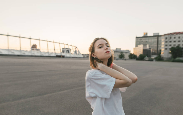 Beautiful girl in a white T-shirt poses background of a city landscape at dawn,looks in camera and holds his hands on the neck.Street portrait of a stylish girl in casual clothing on an evening walk. - 写真・画像