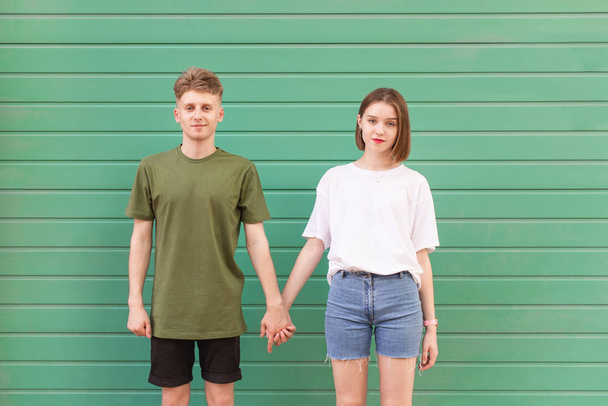 Mladý muž a atraktivní dívka se drží za ruce na tyrkysově pozadí a dívejte se do kamery. Krásný mladý pár stojící na pozadí zelené zdi a pózný na kameře - Fotografie, Obrázek