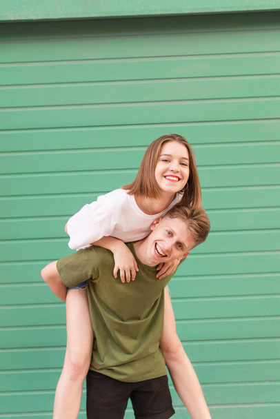 Счастливая пара на фоне цветной зеленой стены, мальчик держит девушку на спине, смотрит в камеру и улыбается. Вертикальное фото. Уличный портрет
. - Фото, изображение