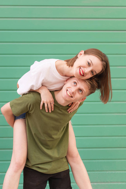 Portrait of a happy couple on a turquoise background, a young man raised the girl on her back, the girl smiles and looks at the camera. Girl hugs a guy, he holds her on her back - Photo, Image