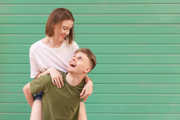 Happy man carries his girlfriend on his back, looking at her and smiling. Portrait of a young lovers couple against the backdrop of a green wall wearing summer casual clothing. - Foto, afbeelding