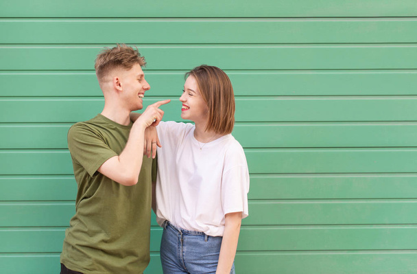 Beautiful young couple guy and girl on green background, looking at each other and smiling. Street portrait of a beautiful young couple in a casual wear against a background of a turquoise wall - Foto, afbeelding