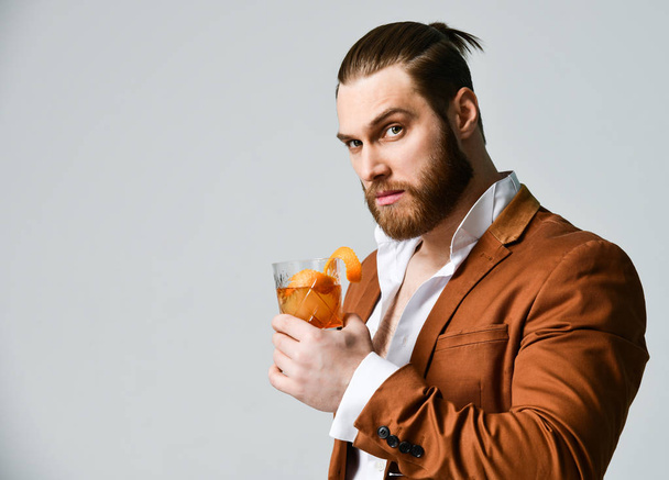 Stylish modern man bearded clubber in fashion suit looks skeptically at the glass with fresh cocktail drink - 写真・画像