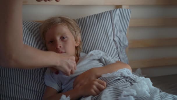 Superslowmotion shot of a sick little boy in a bed. Mother measures his temperature and kisses him. Baby flu concept - Záběry, video