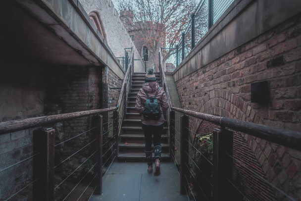 Visão traseira da silhueta de uma jovem caminhando por uma escada entre paredes de tijolos antigos de um castelo antigo em um dia nublado e nebuloso
 - Foto, Imagem