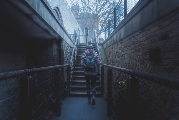 Visão traseira da silhueta de uma jovem caminhando por uma escada entre paredes de tijolos antigos de um castelo antigo em um dia nublado e nebuloso
 - Foto, Imagem