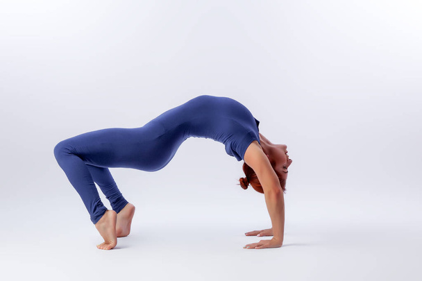 Beautiful slim woman in sports overalls  doing yoga, standing in an asana balancing pose - bridge outside of an overt bow on white  isolated background. The concept of sports and meditation. Training for stretching and yoga - Fotó, kép