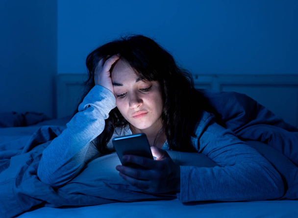 Addicted young woman chatting and surfing on the internet using her smart phone sleepy, bored and tired late at night. Dramatic dark light. In Internet, Mobile addiction and insomnia concept. - Foto, imagen