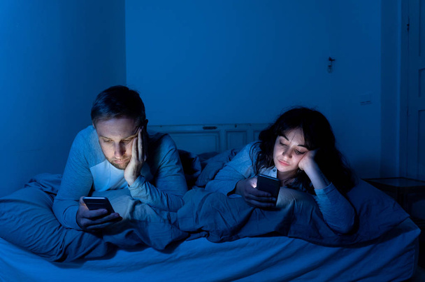 Life style portrait of young bored couple in bed at night on smart phones obsessed with games, social media, apps ignoring each other. Relationship communication problems and phone addiction concept. - Фото, изображение