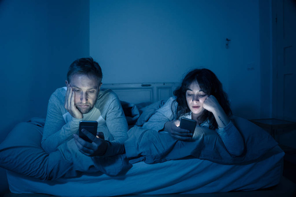 Life style portrait of young bored couple in bed at night on smart phones obsessed with games, social media, apps ignoring each other. Relationship communication problems and phone addiction concept. - Fotografie, Obrázek