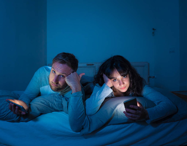 Life style portrait of young bored couple in bed at night on smart phones obsessed with games, social media, apps ignoring each other. Relationship communication problems and phone addiction concept. - 写真・画像
