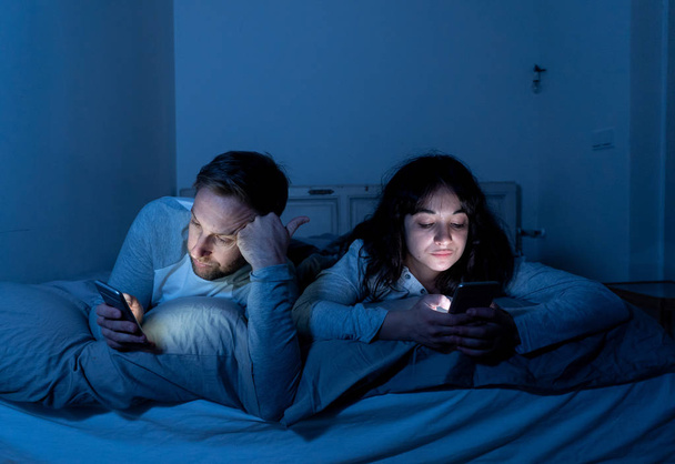 Life style portrait of young bored couple in bed at night on smart phones obsessed with games, social media, apps ignoring each other. Relationship communication problems and phone addiction concept. - Foto, afbeelding