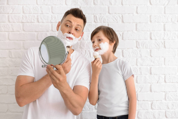 Father and his little son with shaving foam on faces against white brick wall - Photo, Image