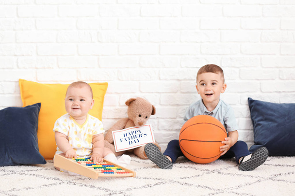 Lindos niños pequeños con juguetes y tarjeta de felicitación para el Día del Padre cerca de la pared de ladrillo blanco
 - Foto, imagen