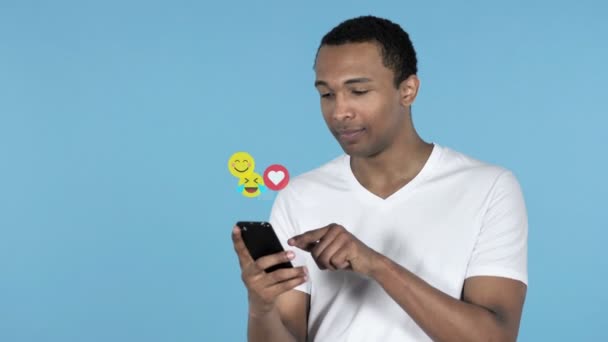 Afričan používá telefon Smartphone izolovaný na modrém pozadí, létající Emotipoje, Emojis a má rád - Záběry, video