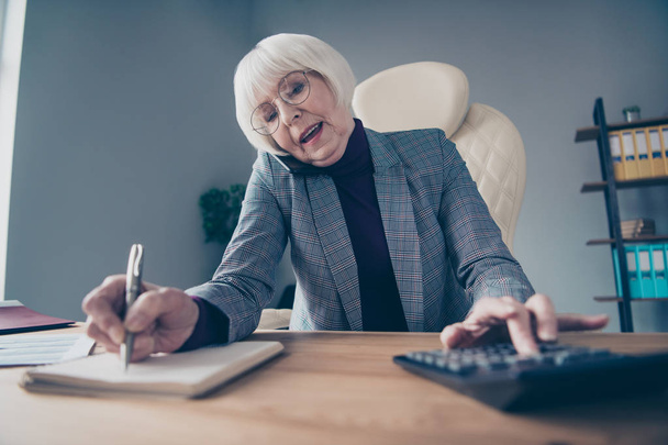 Closeup photo of aged business lady conversation telephone investors noting numbers calculator sit chair wear specs costume jacket - Foto, immagini