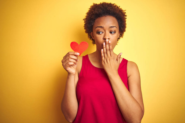 Mujer afroamericana sosteniendo corazones románticos de papel sobre fondo amarillo aislado cubrir la boca con la mano conmocionada con la vergüenza por error, expresión de miedo, asustado en silencio, concepto secreto
 - Foto, imagen