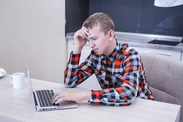 smutný mladý muž v kostlivé košili, který sedí u stolu před notebookem sám. nudný teenager s pohledem na počítač. - Fotografie, Obrázek