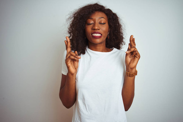 Молода афроамериканець жінка носить футболку стоячи над ізольованою білою тлі жестами пальцем схрещені посміхаючись надією і закритими очима. Концепція удачі і забобонні. - Фото, зображення