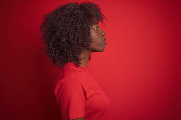 Νεαρή αφρικανική άφρο γυναίκα φορώντας t-shirt στέκεται πάνω από απομονωμένο κόκκινο φόντο κοιτάζοντας προς τα πλάγια, χαλαρώστε το προφίλ πόζα με φυσικό πρόσωπο με αυτοπεποίθηση χαμόγελο. - Φωτογραφία, εικόνα