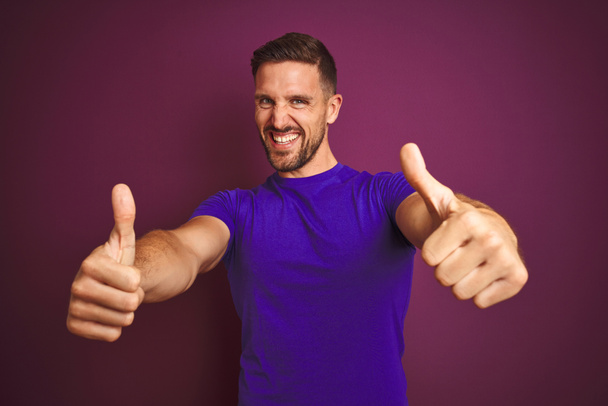 Mladý muž, oblečený v neformálním, purpurovém tričku nad fialovým oblečkem, schvaluje pozitivní gesto rukou, s úsměvem a radostí na úspěch. Gesto vítězů. - Fotografie, Obrázek